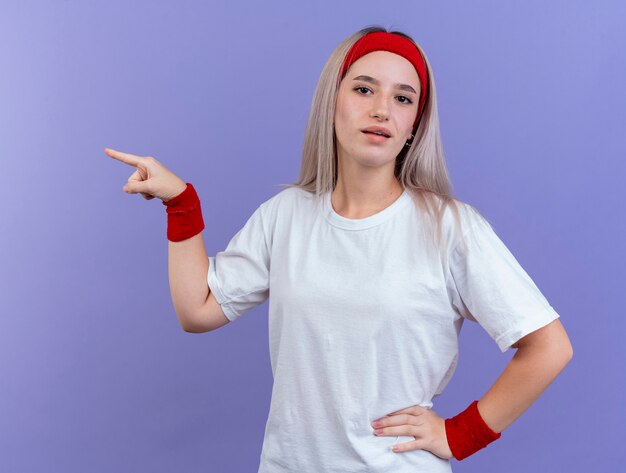 Confident young sporty woman with braces wearing headband and wristbands points at side isolated on purple wall