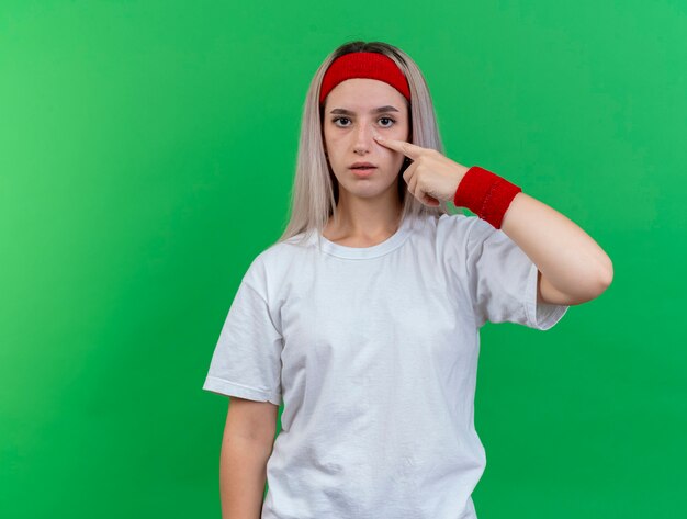 Confident young sporty woman with braces wearing headband and wristbands pulls down eyelid looking at front isolated on green wall