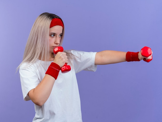 Confident young sporty woman with braces wearing headband and wristbands stands sideways holding dumbbells isolated on purple wall