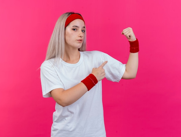 Foto gratuita fiduciosa giovane donna sportiva con le parentesi graffe che indossa la fascia e polsini tempi e punti al bicipite isolato sulla parete rosa