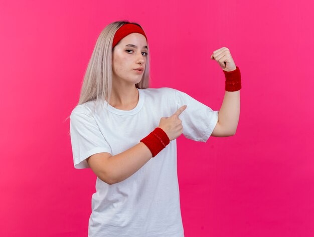 Confident young sporty woman with braces wearing headband and wristbands tenses and points at biceps isolated on pink wall