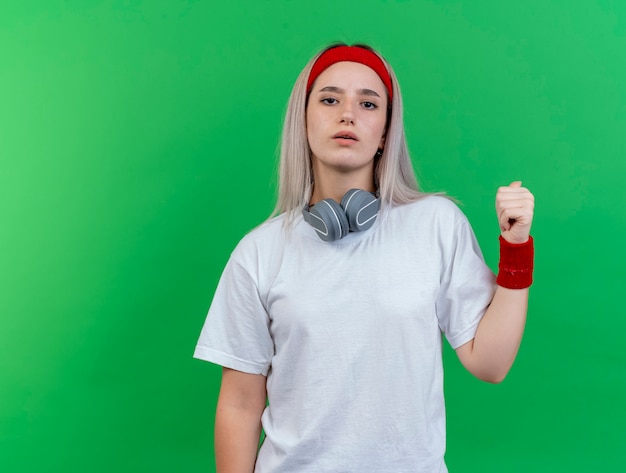 Free photo confident young sporty woman with braces wearing headband and wristbands with headphones on neck points back isolated on green wall