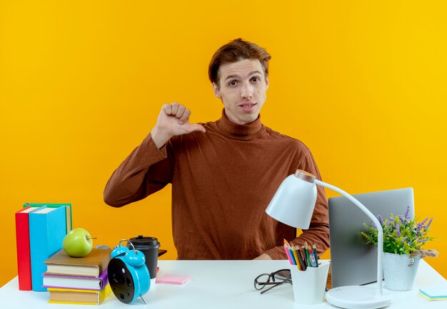 confident young student boy sitting at desk with school tools points at himself