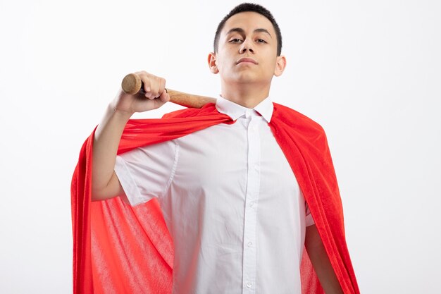 Confident young superhero boy in red cape holding baseball bat on shoulder looking at camera isolated on white background