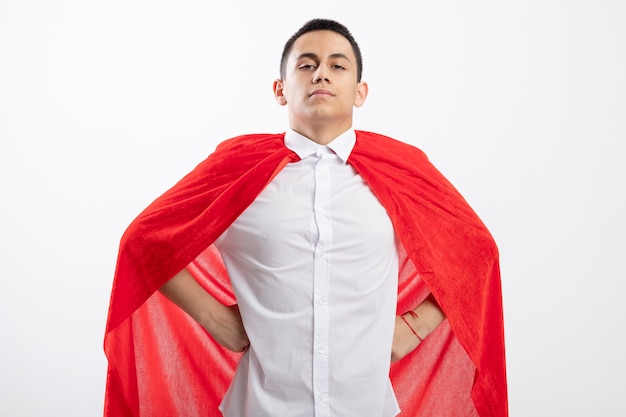 Free photo confident young superhero boy in red cape keeping hands on waist looking at camera isolated on white background