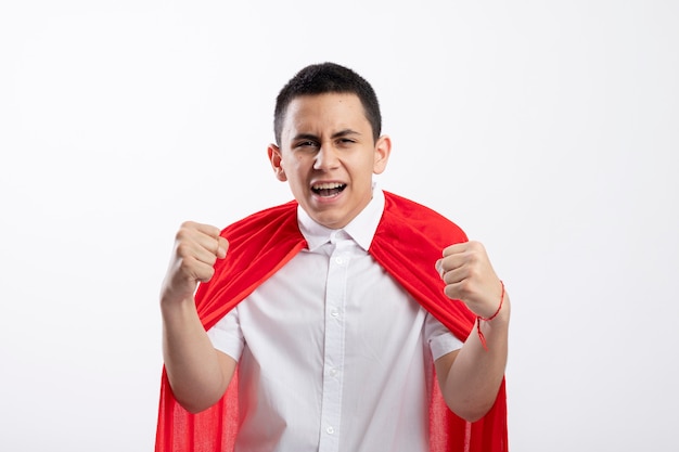 Confident young superhero boy in red cape looking at camera clenching fists isolated on white background with copy space