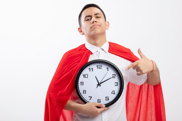 Confident young superhero boy in red cape looking at camera holding and pointing at clock isolated on white background with copy space