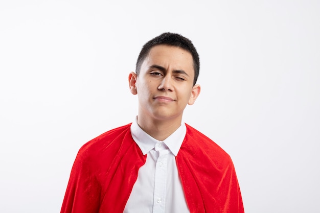 Confident young superhero boy in red cape looking at camera and winking isolated on white background with copy space