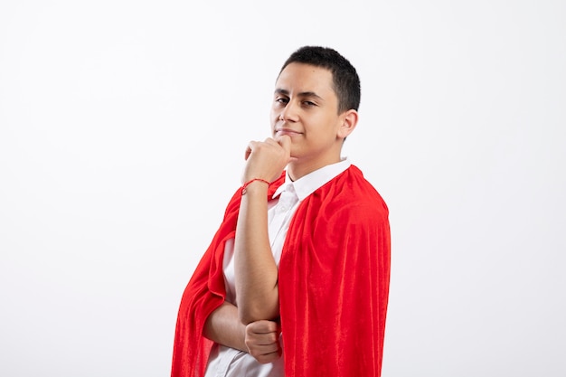 Confident young superhero boy in red cape standing in profile view touching chin looking at camera isolated on white background with copy space