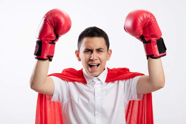 Confident young superhero boy in red cape wearing box gloves looking at camera raising fists up screaming isolated on white background