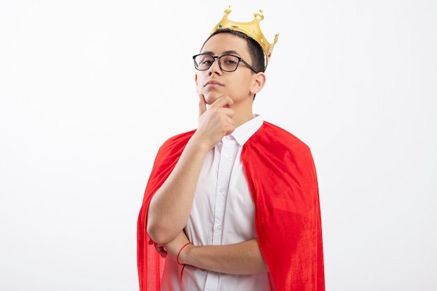 Confident young superhero boy in red cape wearing glasses and crown looking at camera touching chin isolated on white background with copy space