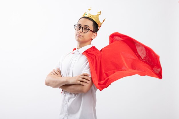 Confident young superhero boy in red cape wearing glasses and crown standing with closed posture in profile view looking at camera isolated on white background with copy space
