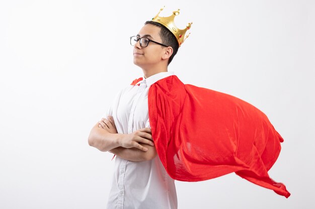 Confident young superhero boy in red cape wearing glasses and crown standing with closed posture in profile view looking straight isolated on white background with copy space