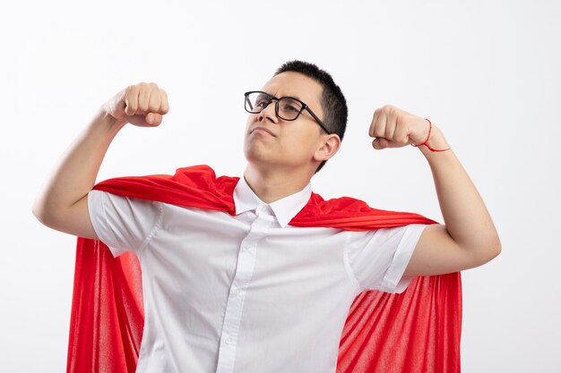 Confident young superhero boy in red cape wearing glasses doing strong gesture looking at side isolated on white background