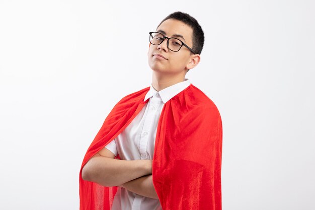 Confident young superhero boy in red cape wearing glasses standing with closed posture in profile view isolated on white background with copy space