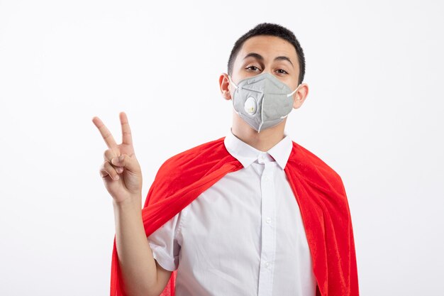 Confident young superhero boy in red cape wearing protective mask doing peace sign looking at camera isolated on white background