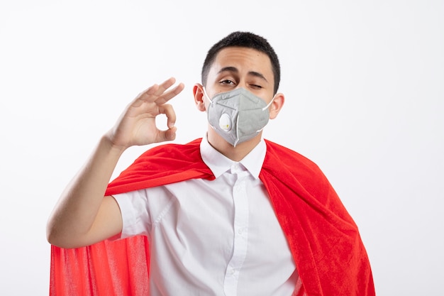 Confident young superhero boy in red cape wearing protective mask looking at camera doing ok sign isolated on white background