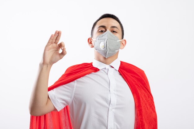 Confident young superhero boy in red cape wearing protective mask looking at camera doing ok sign isolated on white background