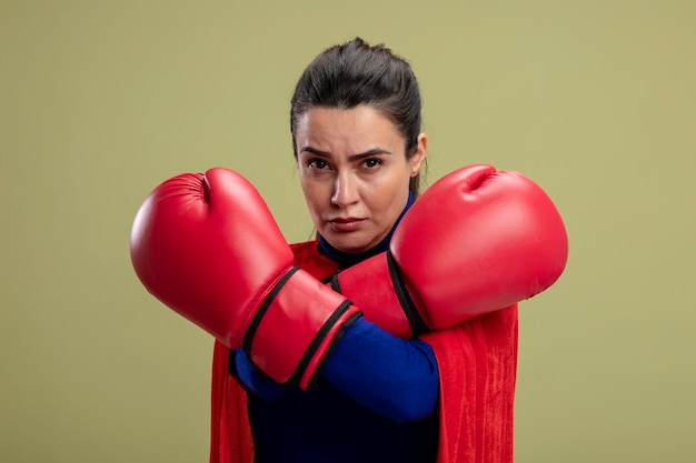 Confident young superhero girl wearing boxing gloves crossing hands isolated on olive green background