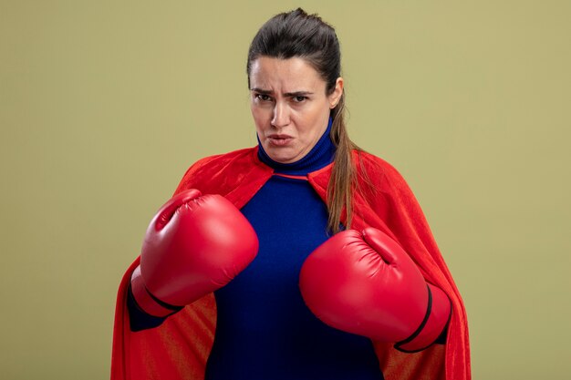 Confident young superhero girl wearing boxing gloves standing in fighting pose isolated on olive green background