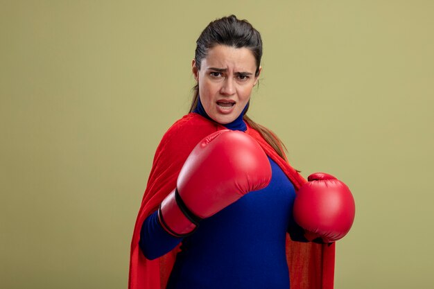 Confident young superhero girl wearing boxing gloves standing in fighting pose isolated on olive green