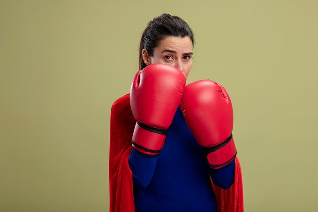 Confident young superhero girl wearing boxing gloves standing in fighting pose isolated on olive green