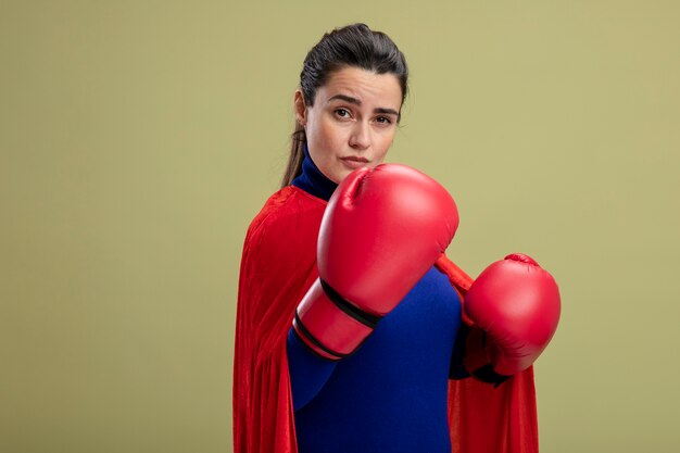 Confident young superhero girl wearing boxing gloves standing in fighting pose isolated on olive green