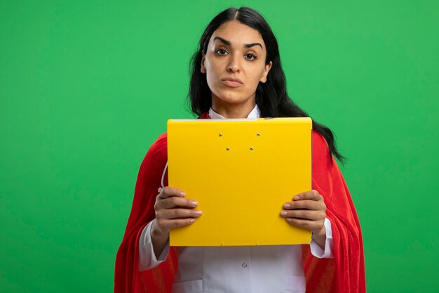 Confident young superhero girl wearing medical robe with stethoscope holding folder isolated on green