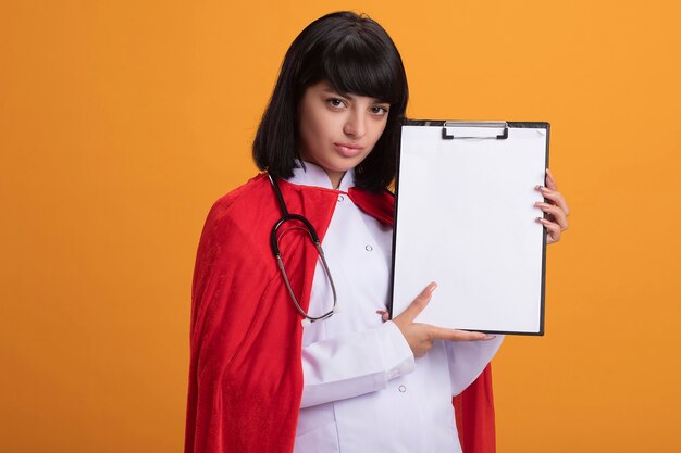 Уверенная молодая девушка супергероя в стетоскопе с медицинским халатом и плащом с буфером обмена