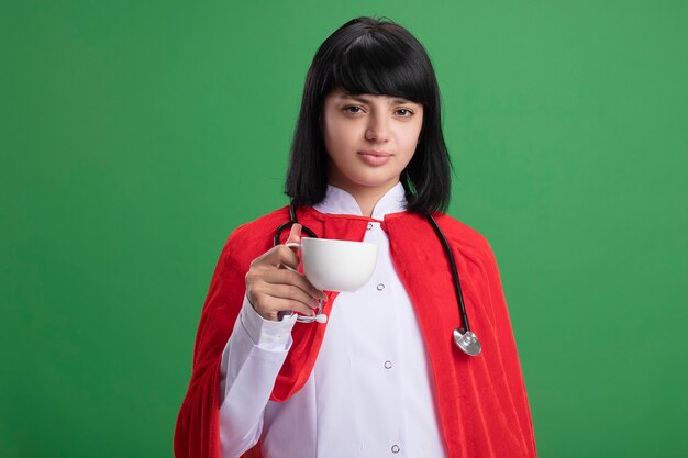 Confident young superhero girl wearing stethoscope with medical robe and cloak holding cup of tea isolated on green