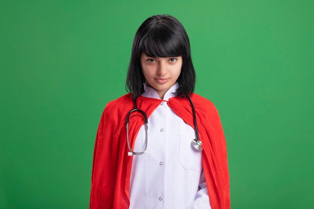 Confident young superhero girl wearing stethoscope with medical robe and cloak isolated on green