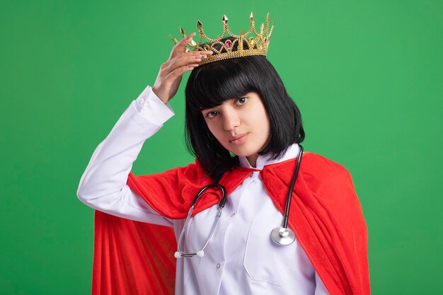 Confident young superhero girl wearing stethoscope with medical robe and cloak putting crown on head isolated on green