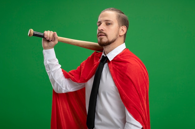 Confident young superhero guy holding baseball bat on shoulder isolated on green background