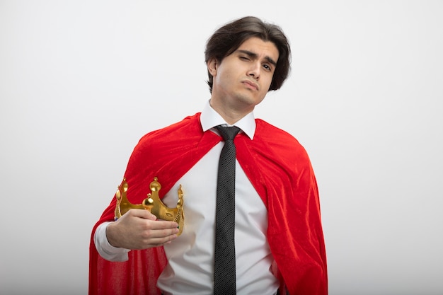 Confident young superhero guy looking at camera wearing tie holding crown isolated on white background