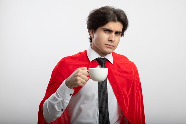 Confident young superhero guy looking at camera wearing tie holding cup of coffee isolated on white