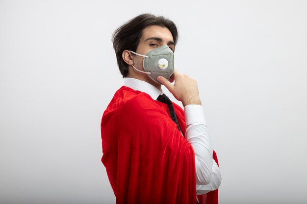 Confident young superhero guy looking at camera wearing tie and medical mask putting hand on chin