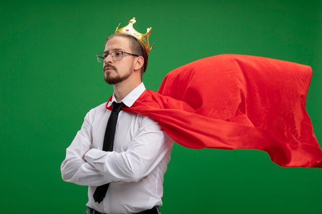 Confident young superhero guy looking at side wearing crown and tie crossing hands isolated on green