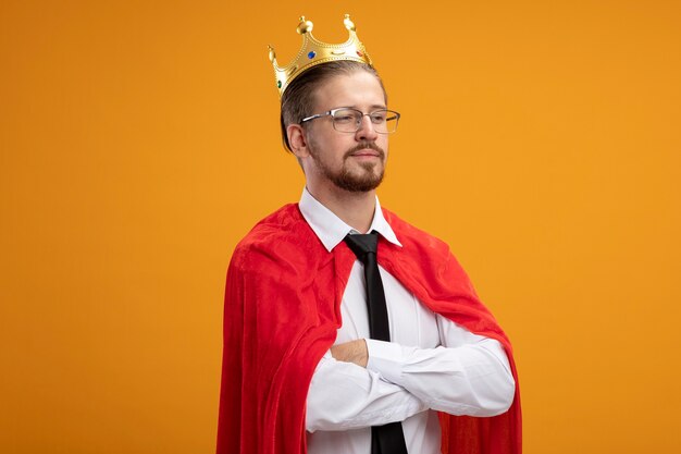 Confident young superhero guy looking at side wearing tie and crown with glasses crossing hands isolated on orange background