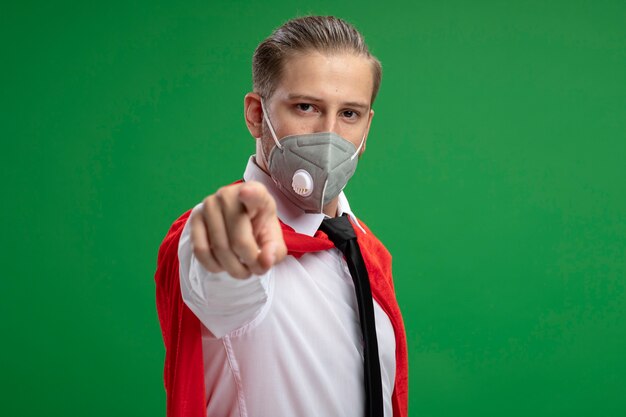 緑の背景に分離されたジェスチャーを示す医療マスクとネクタイを身に着けている自信を持って若いスーパーヒーローの男