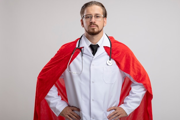 Confident young superhero guy wearing medical robe with stethoscope and glasses putting hands on hip isolated on white background