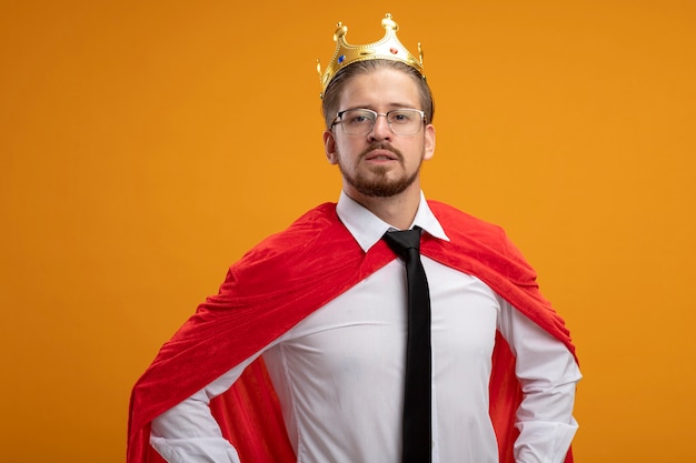 Confident young superhero guy wearing tie and crown with glasses putting hands on hip isolated on orange