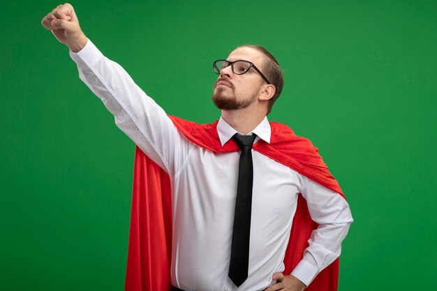 Confident young superhero guy wearing tie and glasses raising fist isolated on green