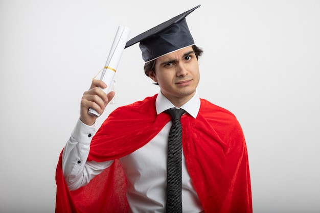 Foto gratuita fiducioso giovane supereroe ragazzo indossa cravatta e cappello laureato innalzamento diploma isolato su sfondo bianco