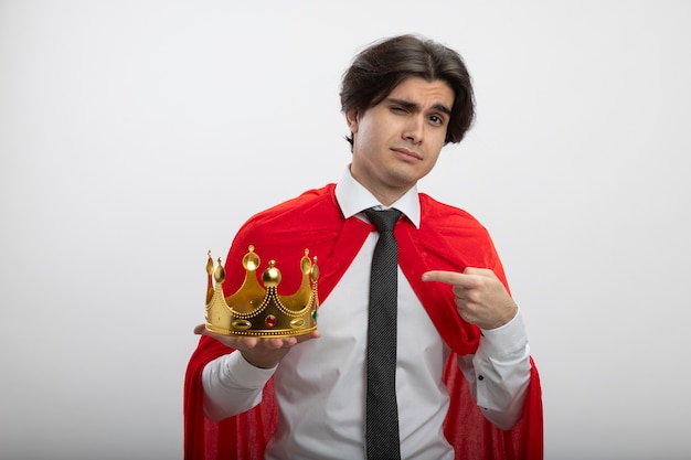 Confident young superhero guy wearing tie holding and points at crown