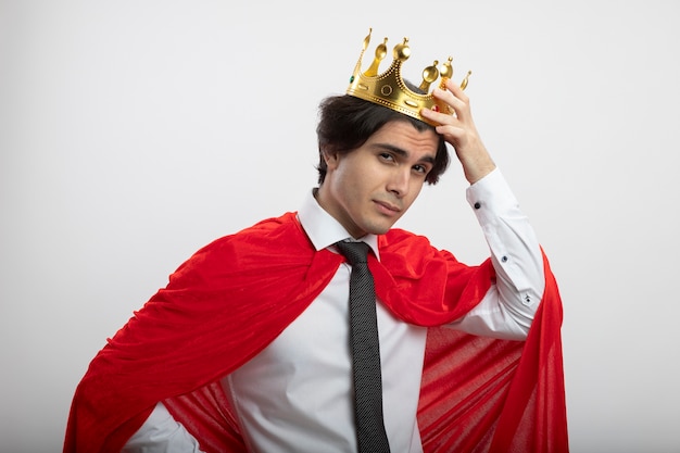 Free photo confident young superhero guy wearing tie putting crown on head isolated on white background