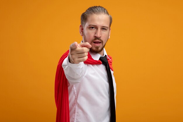 Confident young superhero guy wearing tie showing you gesture isolated on orange background