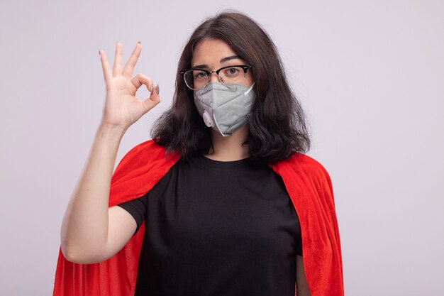 Confident young superhero woman in red cape wearing glasses and protective mask looking at front doing ok sign isolated on white wall