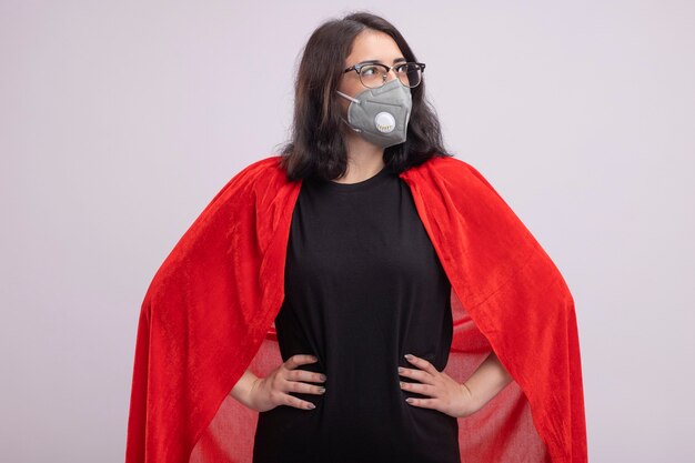 Confident young superhero woman in red cape wearing glasses and protective mask standing like superman looking at side isolated on white wall