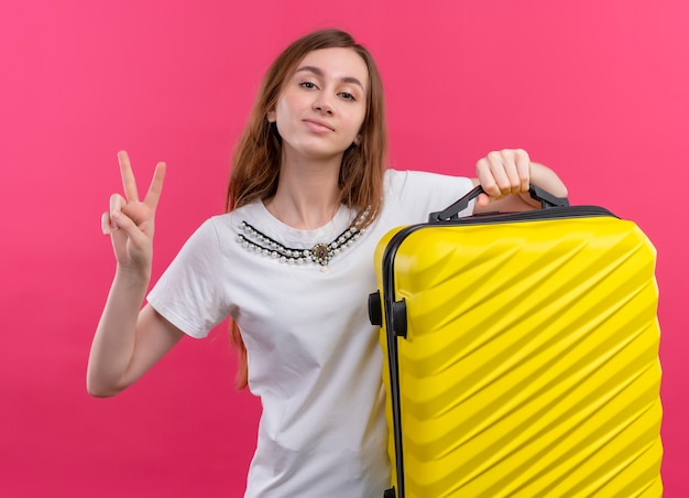 Уверенная молодая девушка-путешественница, держащая чемодан, делает знак мира на изолированном розовом пространстве