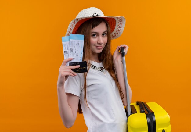 Confident young traveler girl wearing hat holding airplane tickets, credit card and suitcase on isolated orange space with copy space